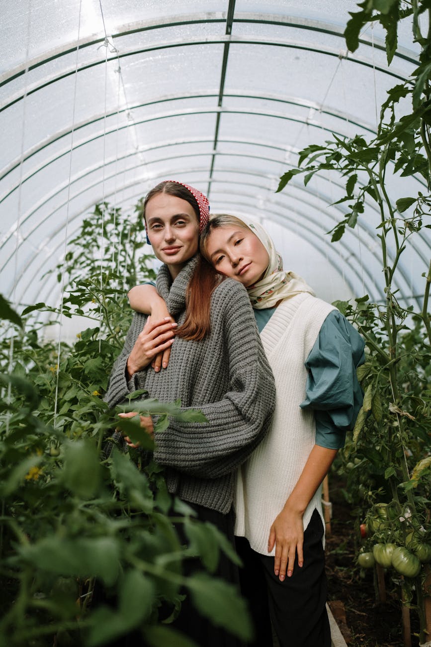 Two young women embrace in a lush greenhouse filled with tomato plants.