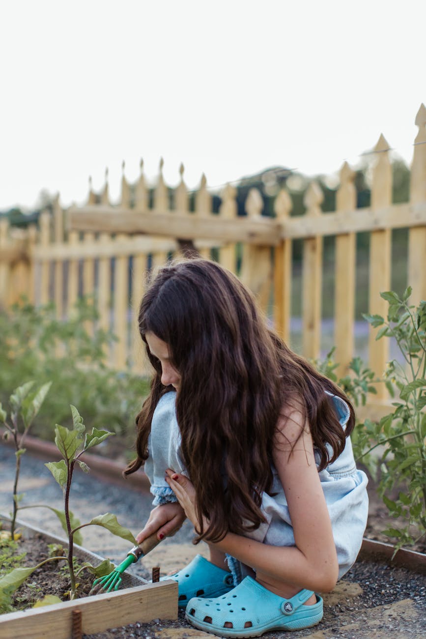A young girl in Crocs uses a gardening fork in an outdoor garden setting.